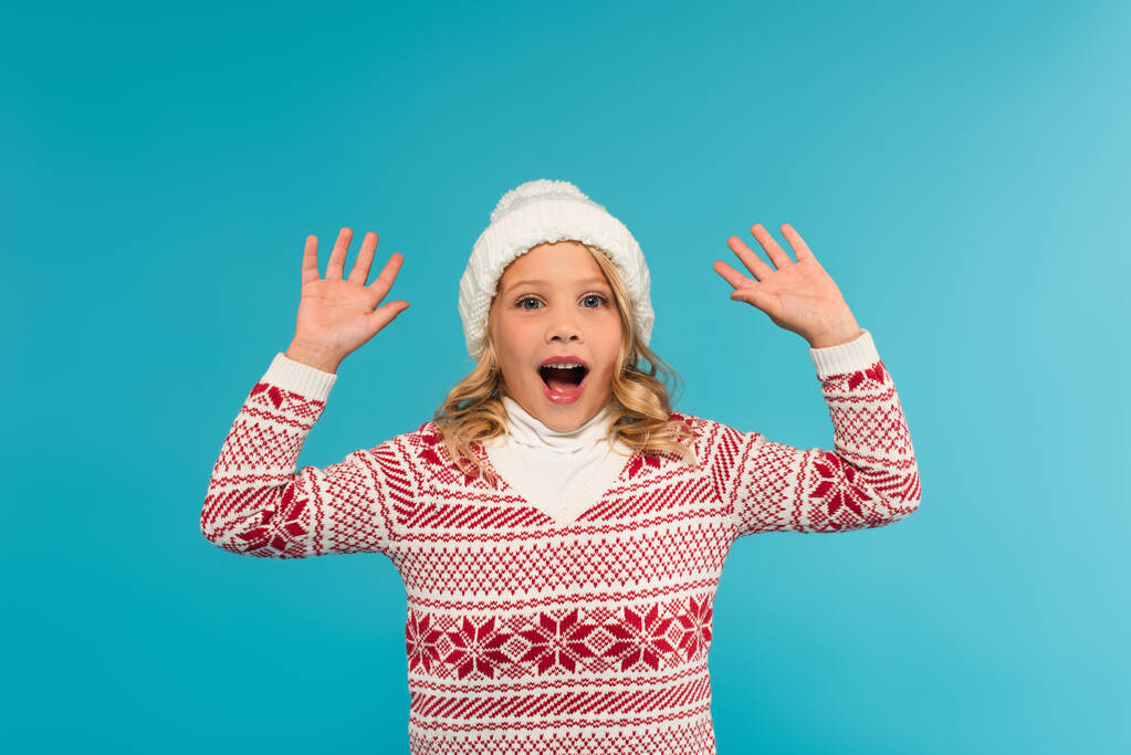взволнованная девушка в теплой шляпе и свитере, показывая вау жест изолирован на синий - Фото, изображение