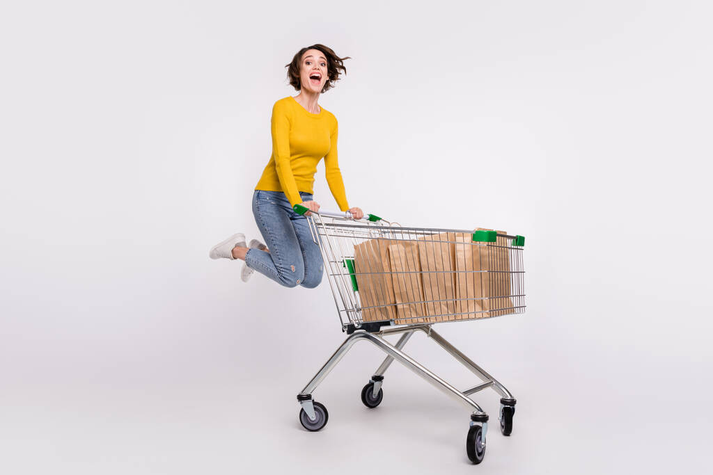 Full length φωτογραφία του αστεία μελαχρινή νεαρή κοπέλα κάνει ψώνια άλμα φορούν τζιν πουκάμισο απομονωμένο σε γκρι φόντο χρώμα - Φωτογραφία, εικόνα