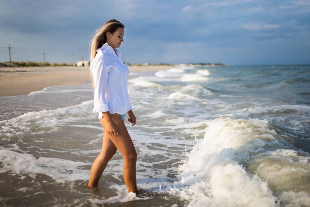 Smukła dziewczyna w delikatnym niebieskim stroju kąpielowym i koszuli spaceruje wzdłuż piaszczystej plaży w pobliżu błękitnego morza - Zdjęcie, obraz