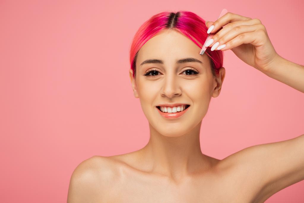 fröhliche junge Frau mit bunten Haaren, die eine Pinzette hält, während sie Augenbrauen isoliert auf rosa modelliert  - Foto, Bild