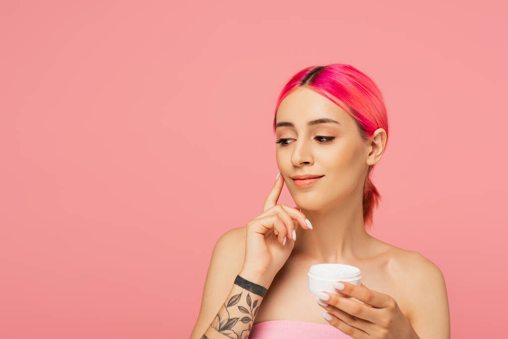 mujer joven tatuada con pelo colorido sonriendo mientras sostiene el recipiente y aplica crema facial aislada en rosa - Foto, imagen