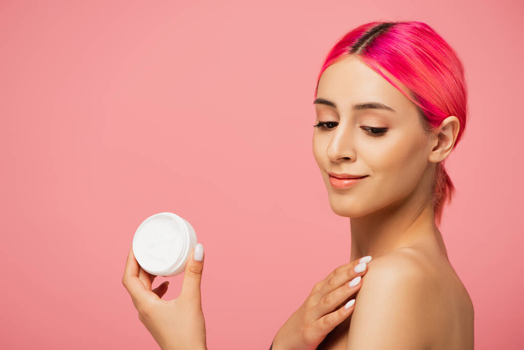 positive junge Frau mit gefärbten Haaren hält Behälter mit kosmetischer Creme und lächelt isoliert auf rosa  - Foto, Bild