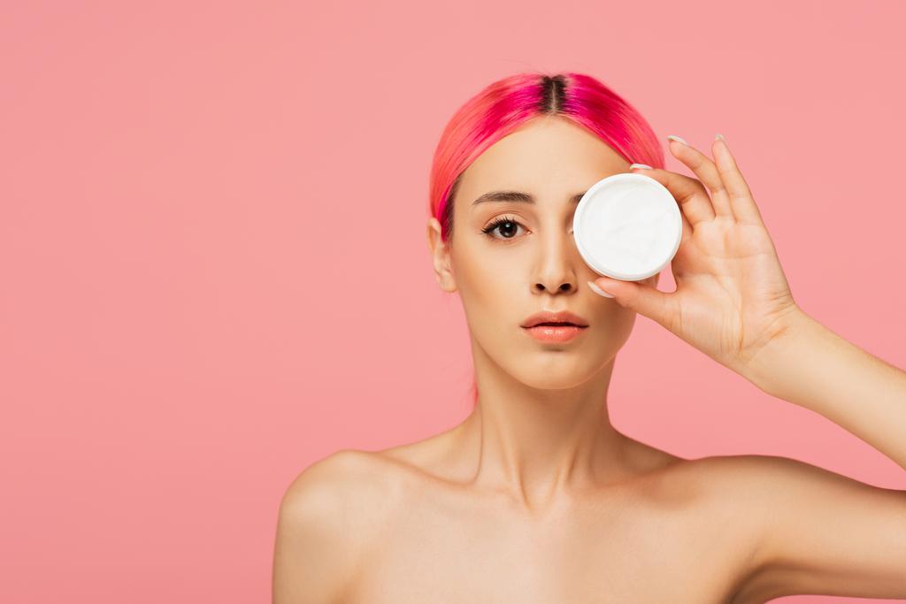young woman with dyed hair covering eye while holding container with cosmetic cream isolated on pink  - Photo, Image