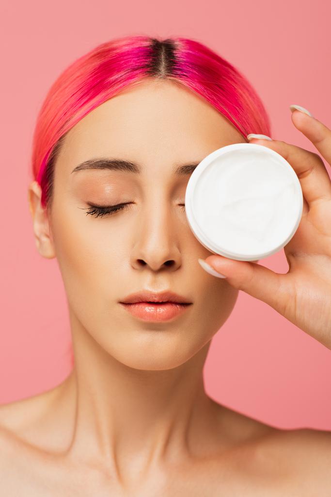 young woman with colorful hair covering eye while holding container with cosmetic cream isolated on pink - Photo, Image