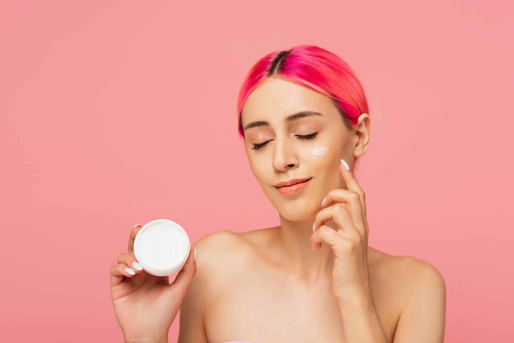 mulher jovem satisfeito com o cabelo tingido segurando recipiente e aplicando creme facial isolado em rosa  - Foto, Imagem