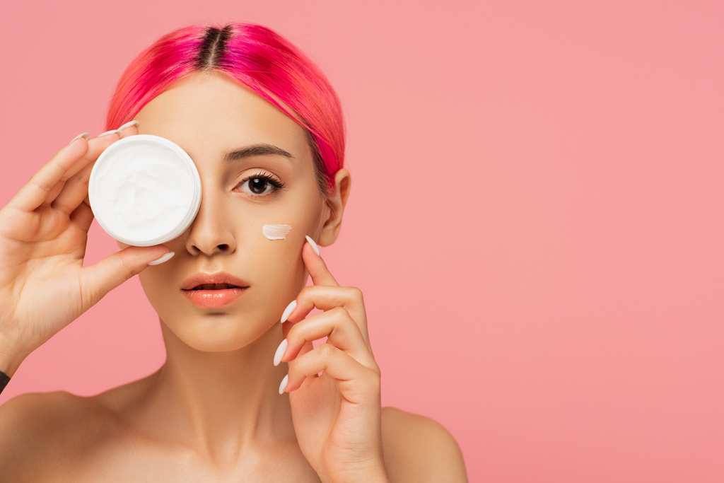 young woman with colorful hair and face cream on cheek holding container while covering eye isolated on pink - Photo, Image