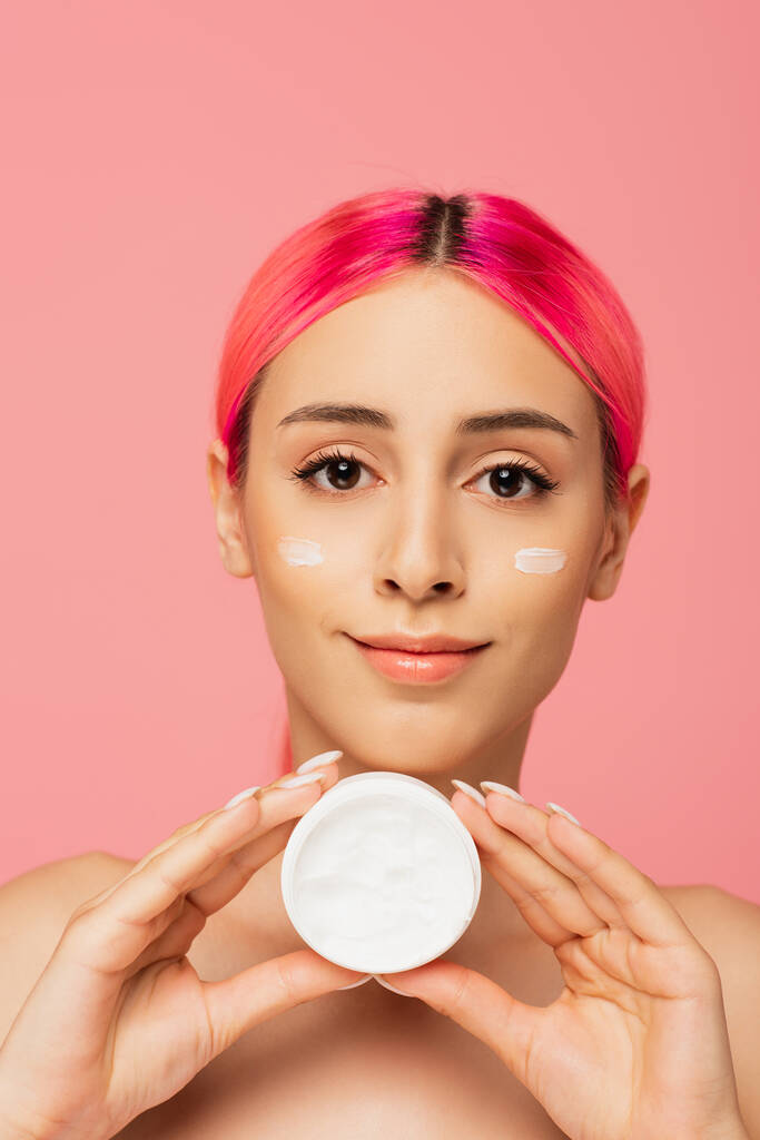 cheerful young woman with colorful hair and cream on cheeks holding container isolated on pink - Photo, Image