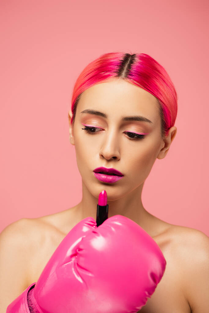 mujer joven con el pelo teñido y maquillaje brillante en guante de boxeo aplicando lápiz labial aislado en rosa  - Foto, imagen