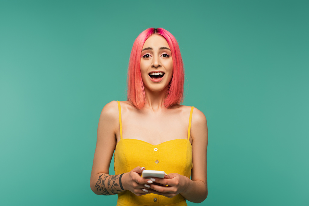 jovem animado com cabelo tingido rosa segurando smartphone isolado em turquesa - Foto, Imagem