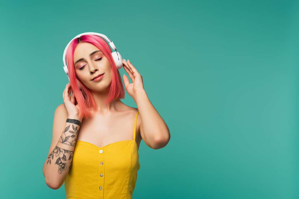 ευχαριστημένη νεαρή γυναίκα με ροζ βαμμένα μαλλιά ακούει μουσική σε ακουστικά που απομονώνονται σε μπλε - Φωτογραφία, εικόνα
