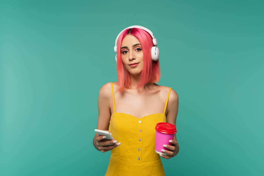 young woman with pink dyed hair listening music in headphones while holding paper cup and smartphone isolated on blue - Photo, Image