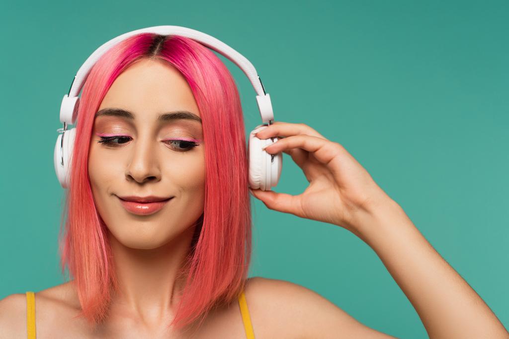 νεαρή γυναίκα με ροζ βαμμένα μαλλιά ακούγοντας μουσική και ρυθμίζοντας ακουστικά που απομονώνονται στο μπλε - Φωτογραφία, εικόνα
