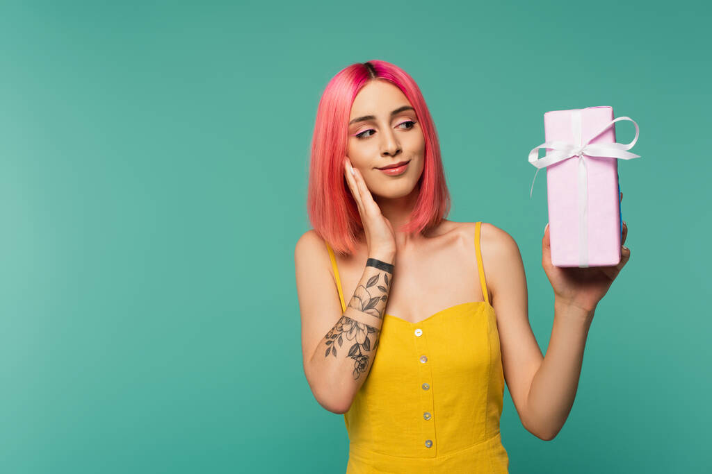 задоволена молода жінка з рожевим фарбованим волоссям тримає загорнутий подарунок ізольовано на синьому
  - Фото, зображення