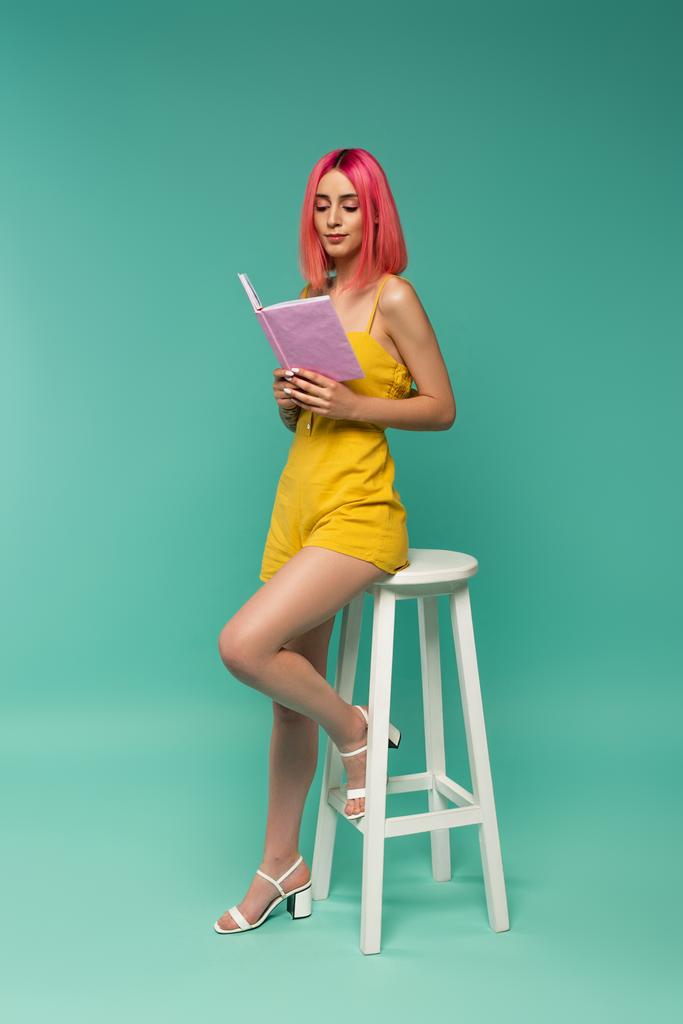 plná délka mladé ženy s růžové barvené vlasy drží knihu, zatímco sedí na stoličce na modré  - Fotografie, Obrázek