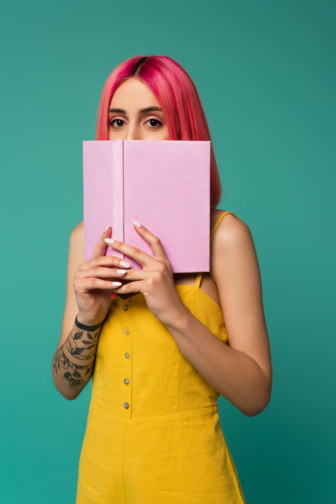 junge Frau mit rosa gefärbten Haaren hält Buch, während sie ihr Gesicht isoliert auf blauem Hintergrund versteckt  - Foto, Bild