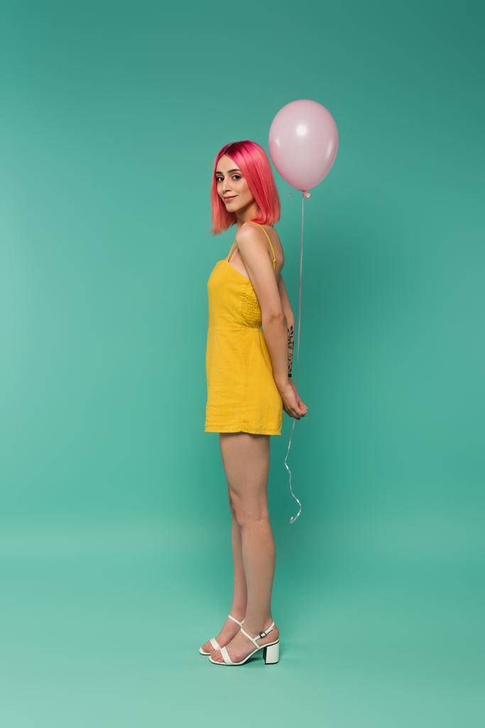 longitud completa de la joven sonriente con el pelo teñido de rosa sosteniendo globo en azul  - Foto, Imagen