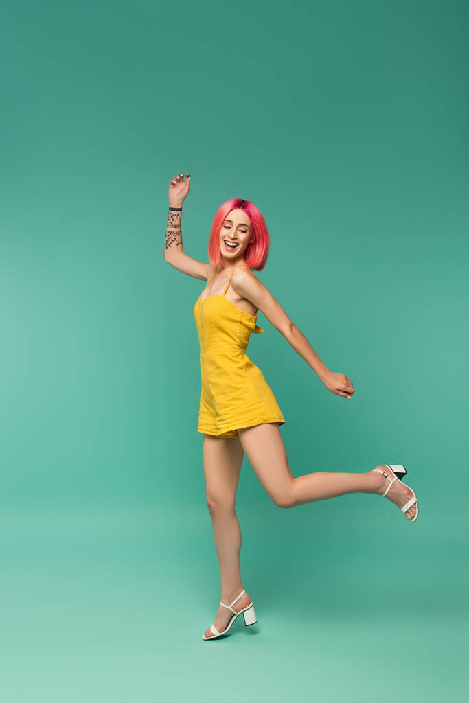 pełna długość uśmiechnięta młoda kobieta z różowymi farbowanymi włosami w romper jumping na niebiesko  - Zdjęcie, obraz