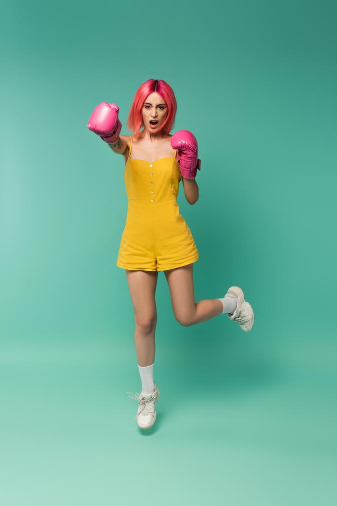 plná délka mladé ženy s růžovými barvenými vlasy v boxerských rukavicích skákání a křik na modré  - Fotografie, Obrázek