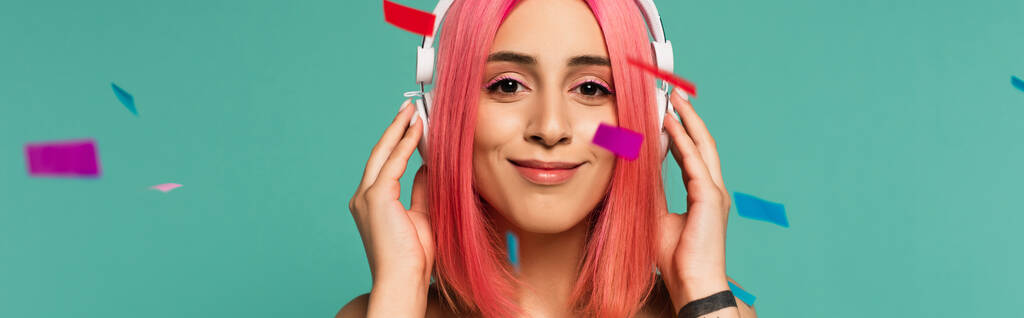 szczęśliwa młoda kobieta z różowymi farbowanymi włosami w bezprzewodowych słuchawkach w pobliżu kolorowego konfetti na niebieskim, baner - Zdjęcie, obraz