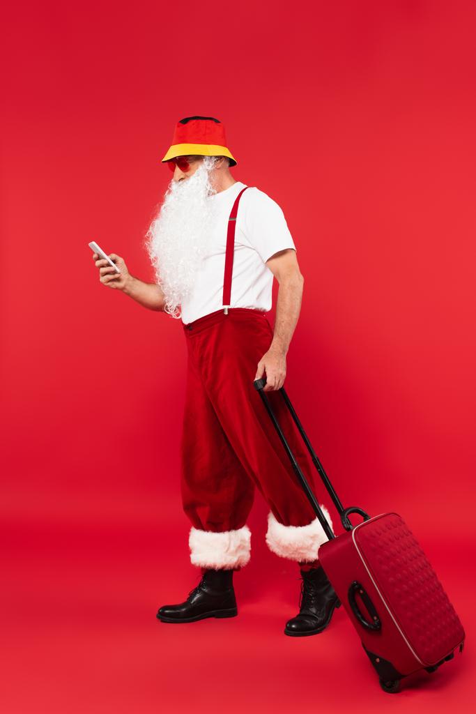Kerstman in zonnebril met behulp van smartphone en het houden van koffer op rode achtergrond - Foto, afbeelding