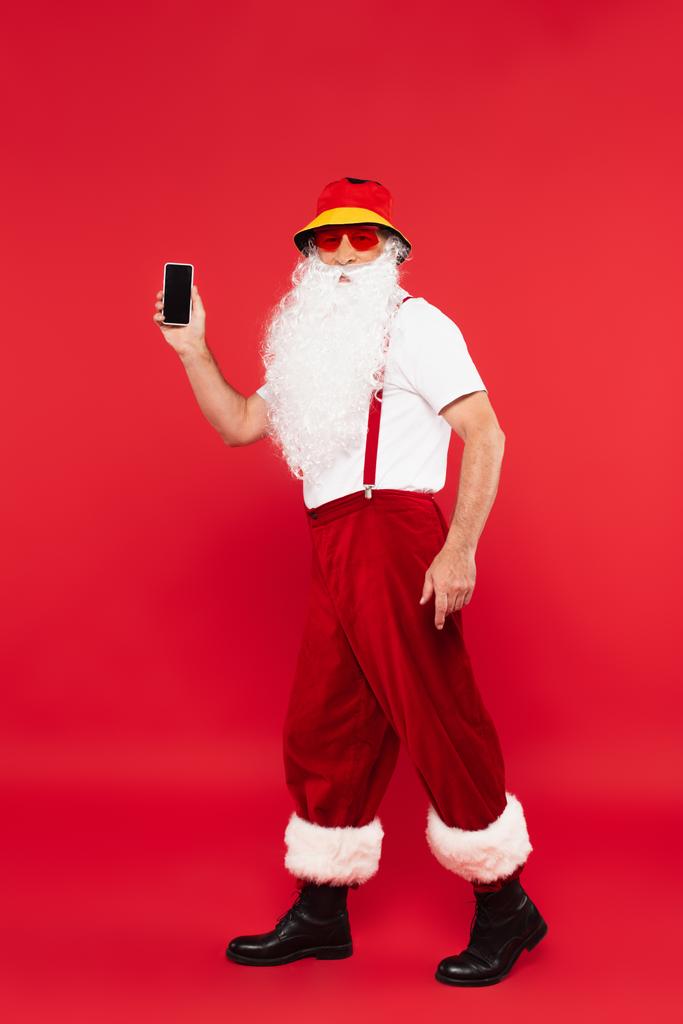 Święty Mikołaj w Panamie i okulary przeciwsłoneczne trzymając smartfona na czerwonym tle - Zdjęcie, obraz