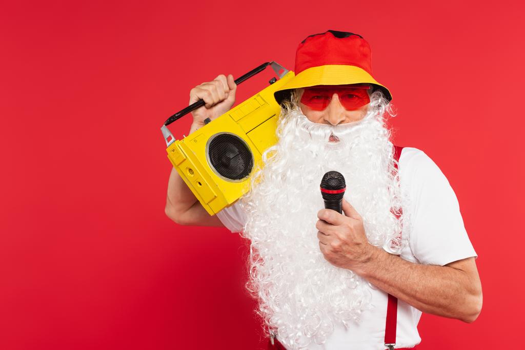 Santa claus in panama and sunglasses holding microphone and boombox isolated on red  - Photo, Image