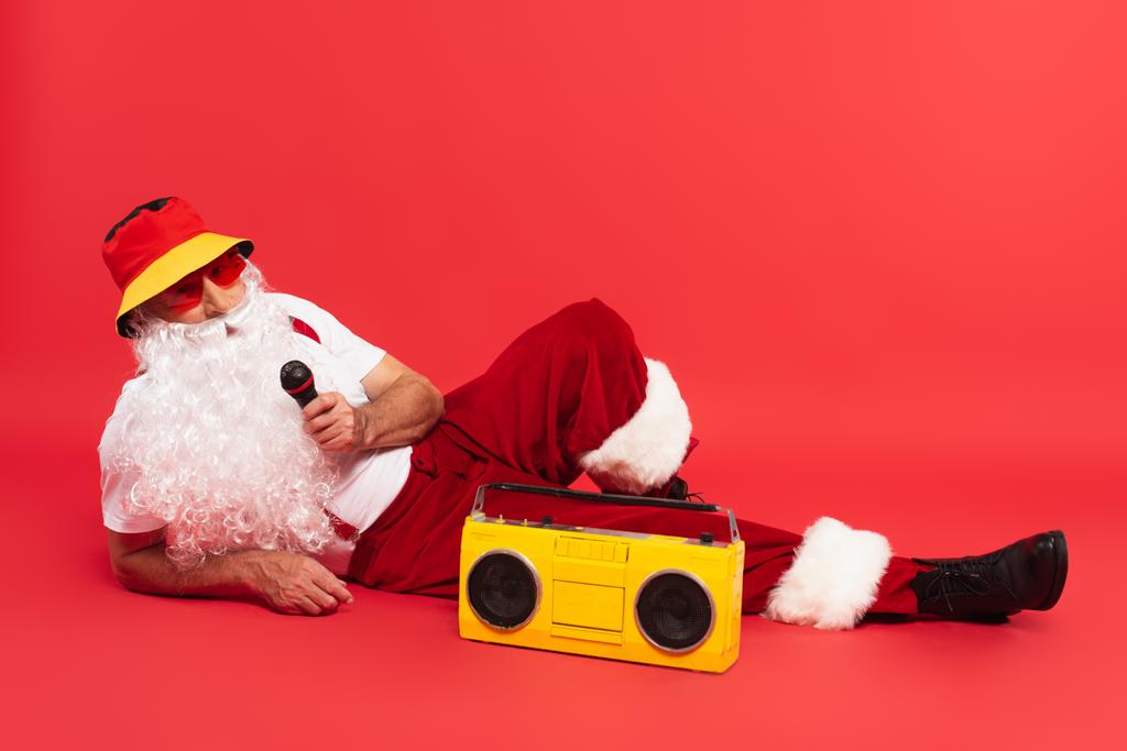 Santa claus in panama holding microphone near boombox on red background - Photo, Image