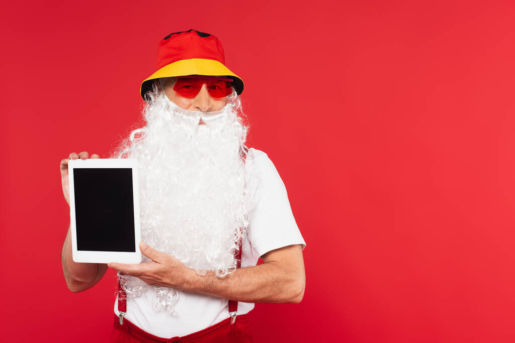 Bärtiger Weihnachtsmann mit Sonnenbrille und digitalem Tablet mit leerem Bildschirm auf rotem Hintergrund  - Foto, Bild