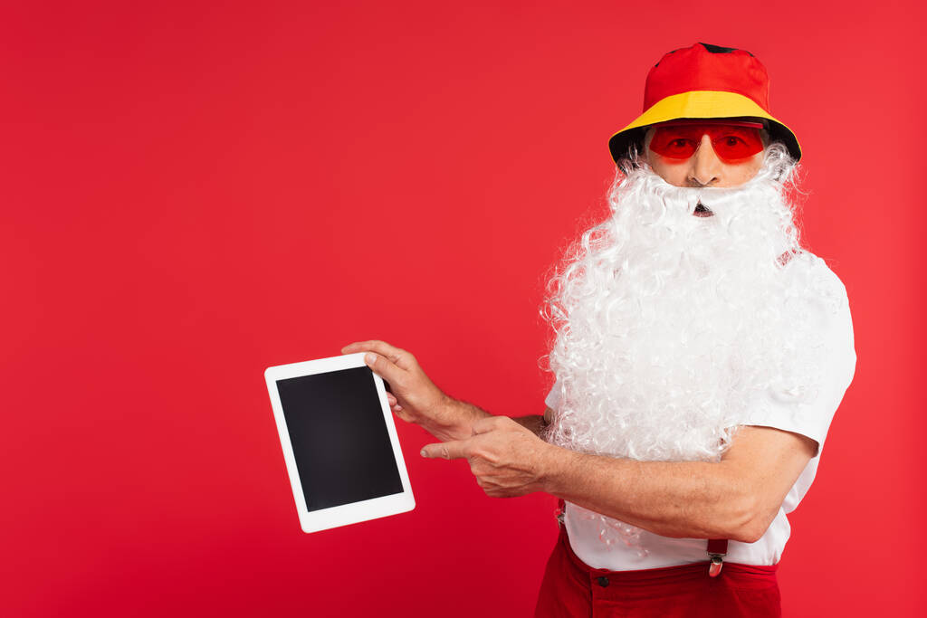 Santa Claus σε panama και γυαλιά ηλίου που δείχνουν σε ψηφιακή ταμπλέτα που απομονώνονται σε κόκκινο  - Φωτογραφία, εικόνα