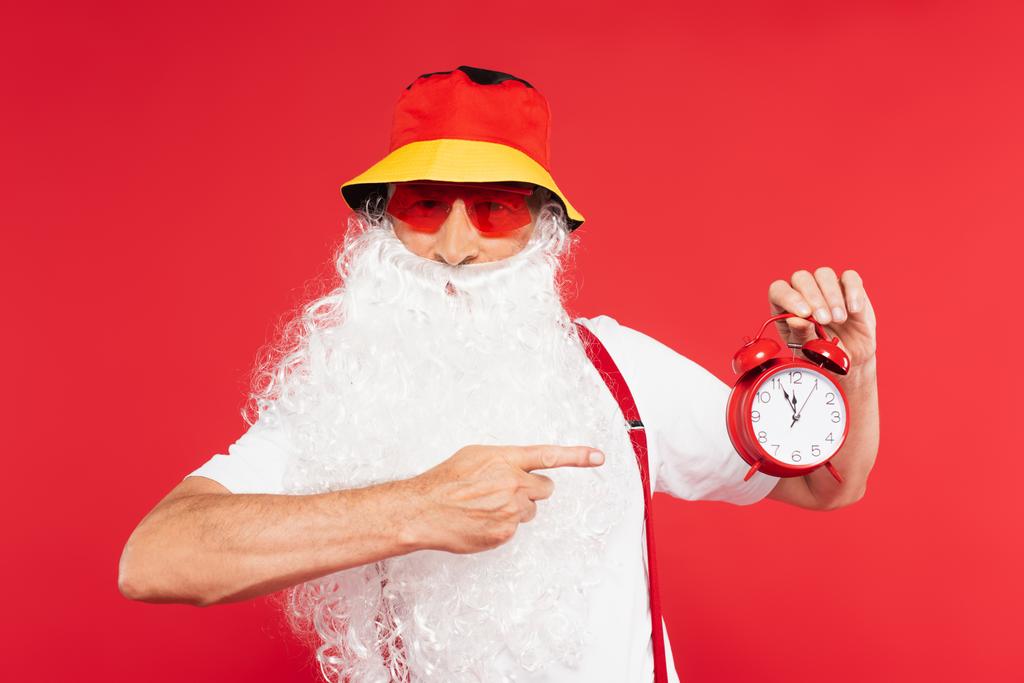 Santa Claus en gafas de sol y panama apuntando al despertador aislado en rojo  - Foto, imagen