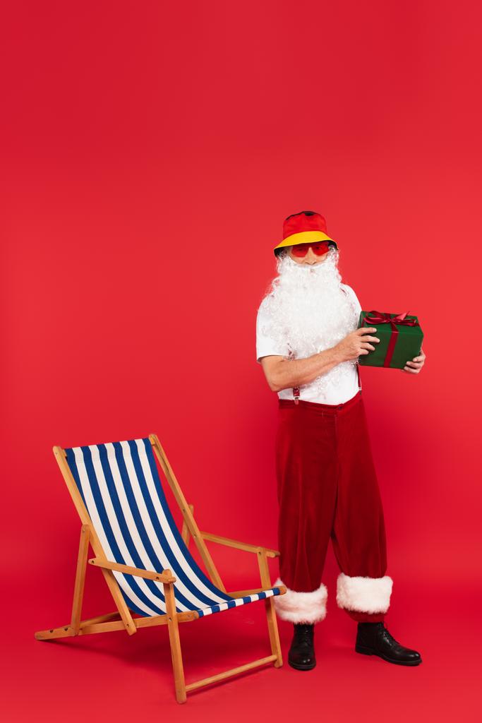 Weihnachtsmann mit Sonnenbrille hält Geschenk neben Liegestuhl auf rotem Hintergrund - Foto, Bild