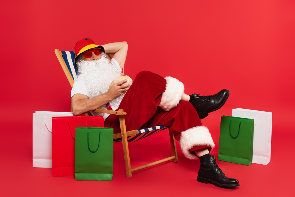 Santa Claus σε παναμά και γυαλιά ηλίου κρατώντας κοκτέιλ στην καρύδα, ενώ κάθεται στην καρέκλα κατάστρωμα κοντά τσάντες ψώνια σε κόκκινο φόντο - Φωτογραφία, εικόνα