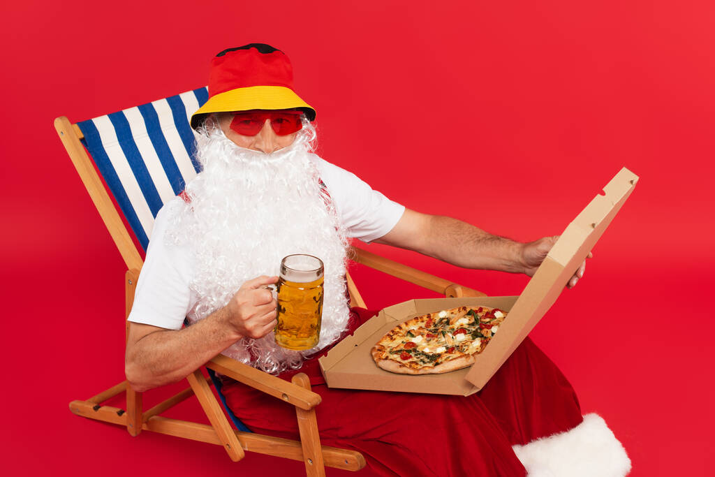 Mikołaj w okularach przeciwsłonecznych z pudełkiem po pizzy i szklanką piwa na leżaku na czerwonym tle - Zdjęcie, obraz