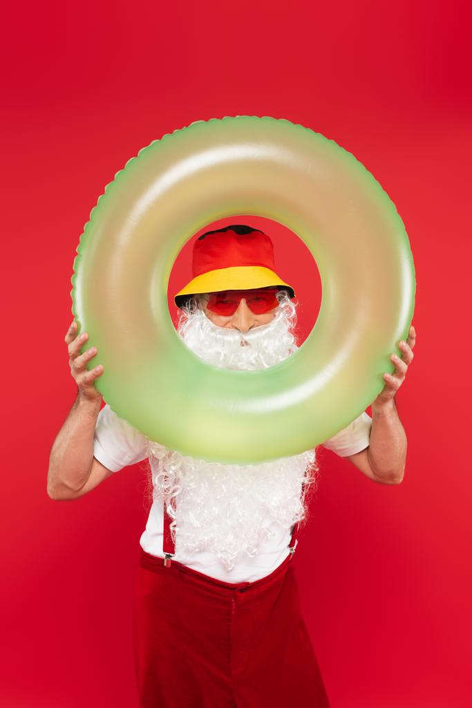 Santa Claus σε Παναμά και γυαλιά ηλίου κοιτάζοντας κάμερα μέσα από δαχτυλίδι κολύμπι απομονώνονται σε κόκκινο  - Φωτογραφία, εικόνα