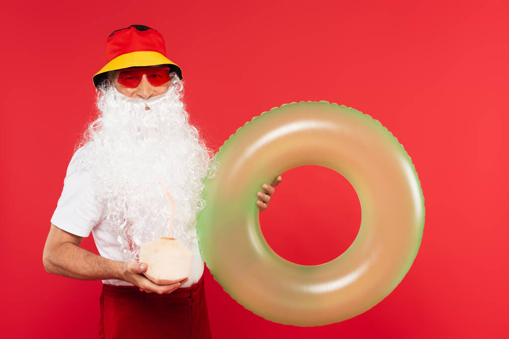 Papai Noel em óculos de sol segurando anel de natação e coquetel em coco isolado em vermelho  - Foto, Imagem
