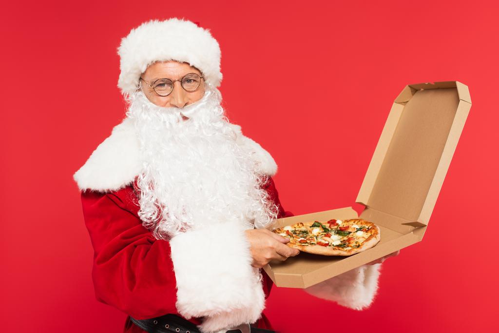 Weihnachtsmann mit Brille und Hut hält Pizza im Karton isoliert auf Rot  - Foto, Bild