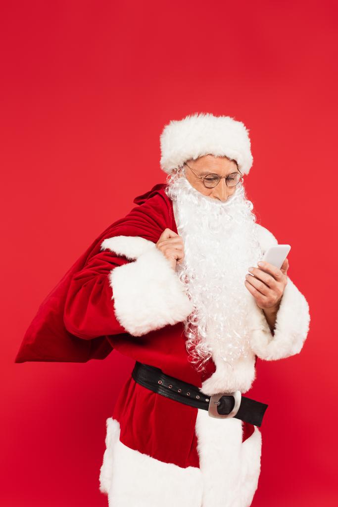 Święty Mikołaj z workiem za pomocą telefonu komórkowego odizolowany na czerwono  - Zdjęcie, obraz