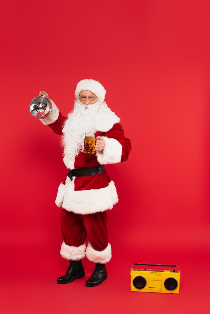 Санта-Клаус с пивом и шаром диско стоит рядом с бумбоксом на красном фоне - Фото, изображение