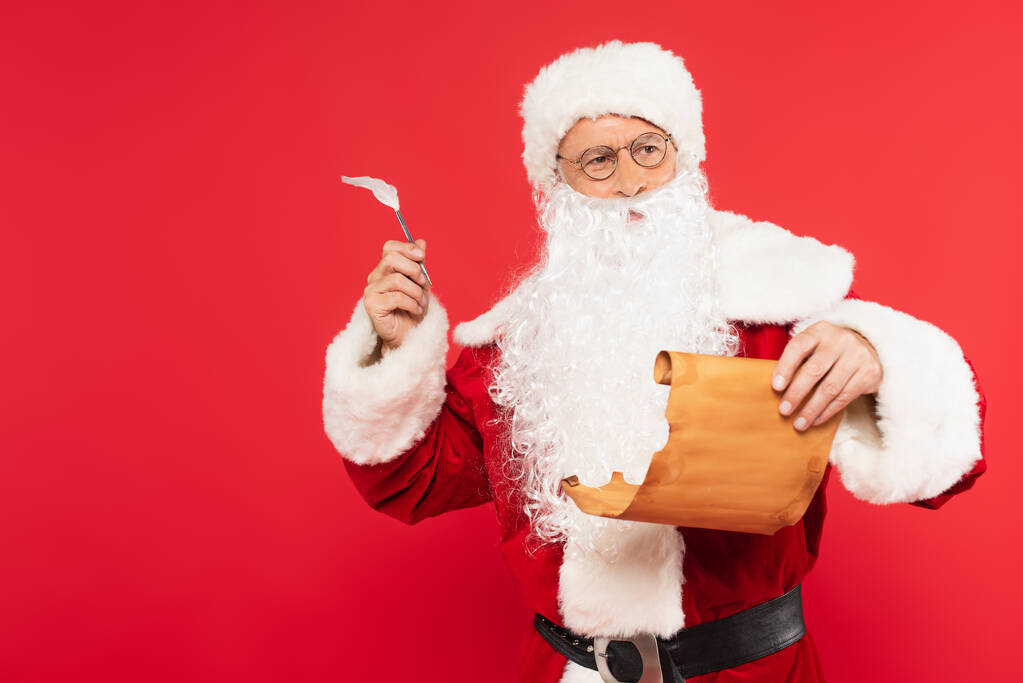 Przemyślany Święty Mikołaj z długopisem i papierem stojącym na czerwonym  - Zdjęcie, obraz