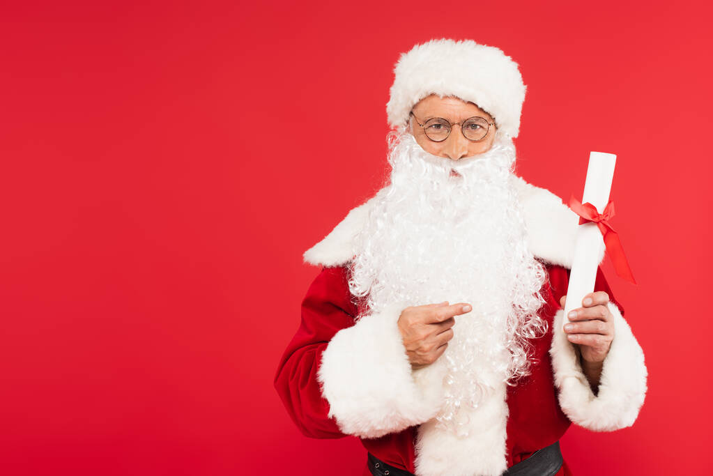 Санта-Клаус указывает на бумагу с луком изолированы на красный  - Фото, изображение