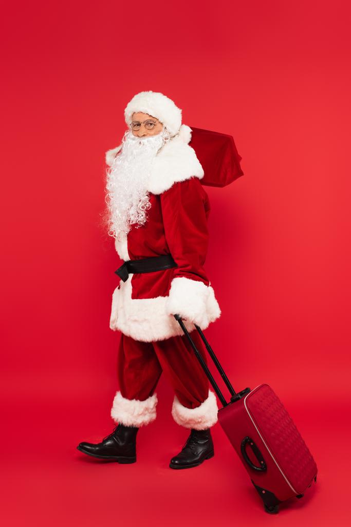 Санта-Клаус в костюмі, що тримає валізу і мішок на червоному тлі
 - Фото, зображення