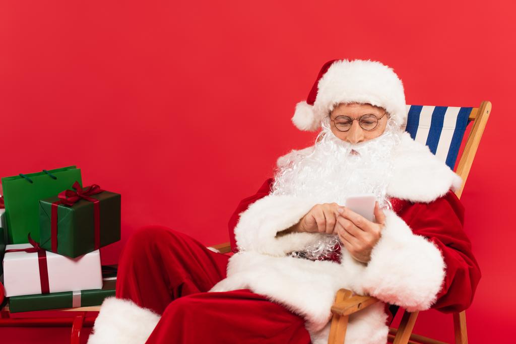Père Noël en utilisant smartphone sur chaise longue près de cadeaux sur traîneau sur fond rouge - Photo, image