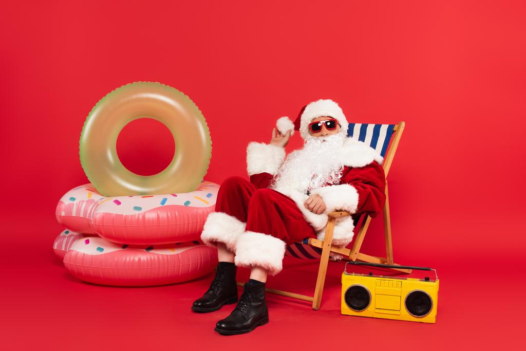 Kerstman in zonnebril en kostuum zitten op dek stoel in de buurt van zwemringen en boombox op rode achtergrond - Foto, afbeelding