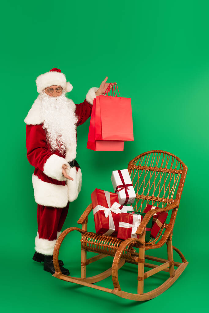 Père Noël en costume tenant des sacs à provisions près de cadeaux sur chaise en osier sur fond vert - Photo, image