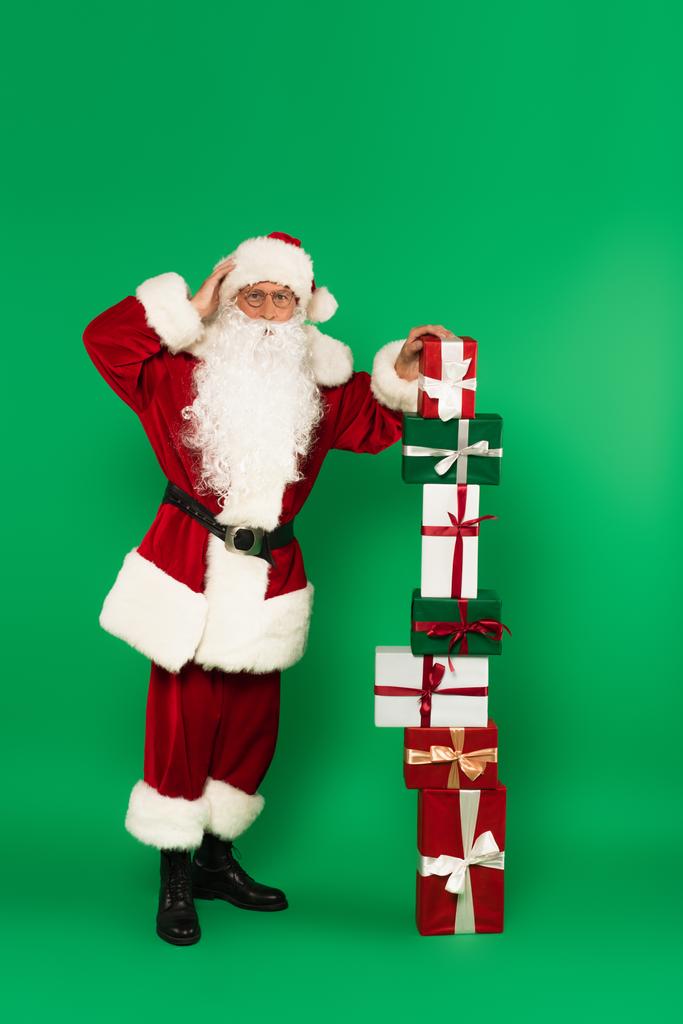 Preocupado Santa Claus de pie cerca de regalos apilados sobre fondo verde - Foto, Imagen
