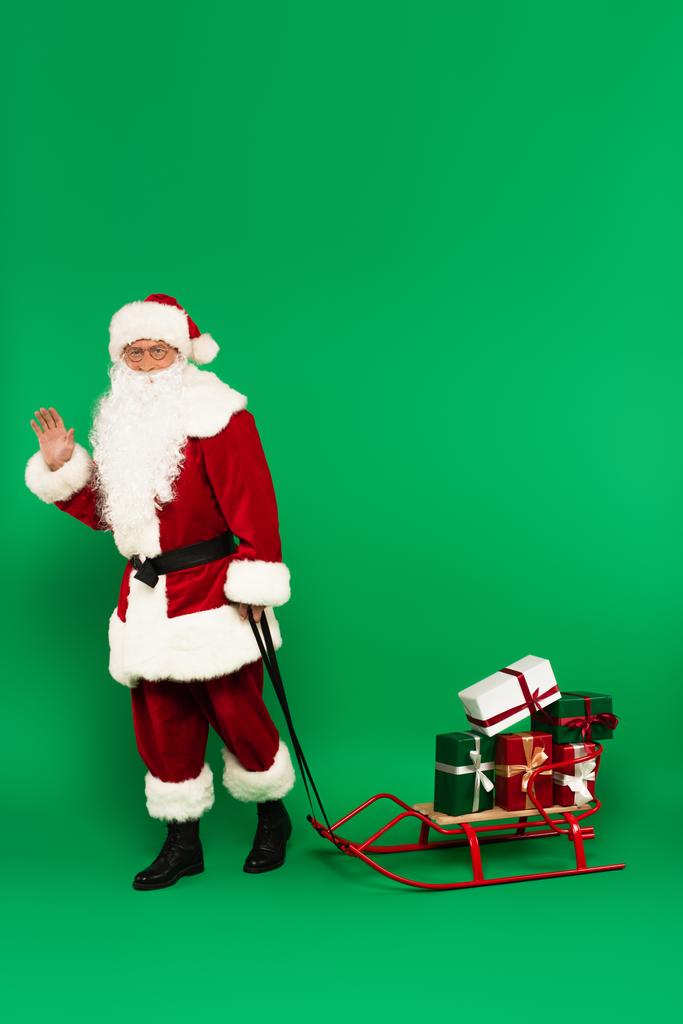 Weihnachtsmann winkt mit der Hand in die Kamera bei Geschenken auf Schlitten auf grünem Hintergrund - Foto, Bild