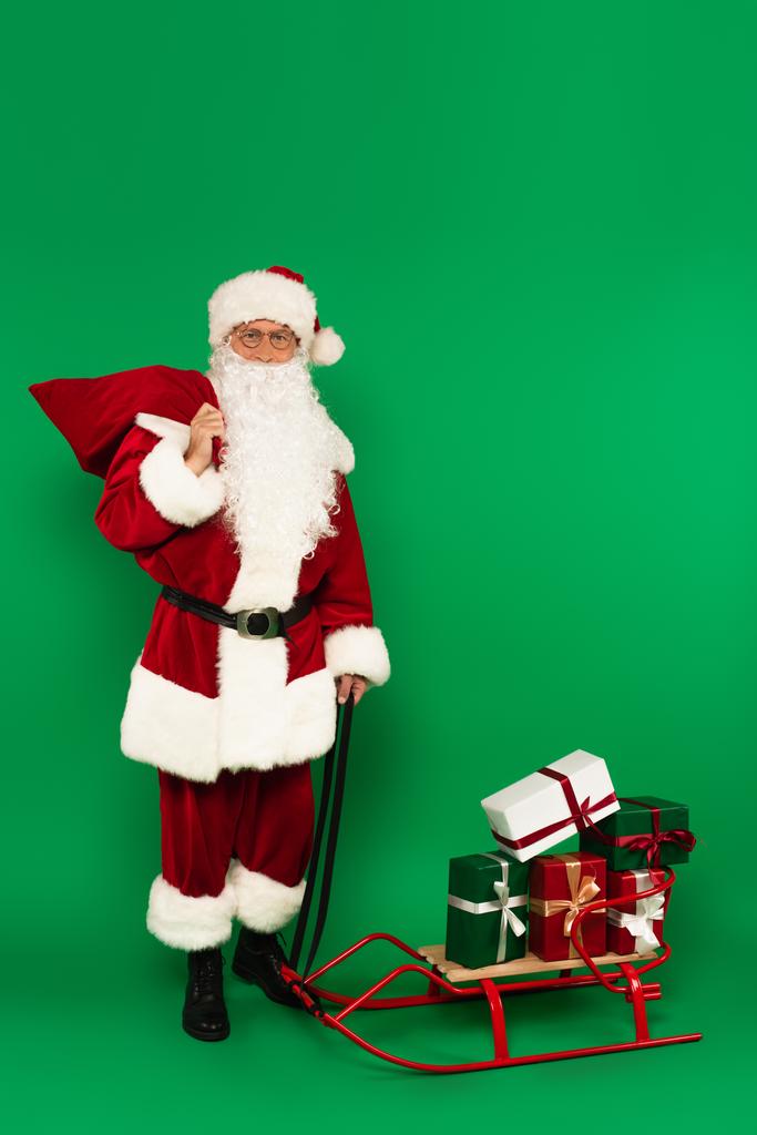 Santa Claus s pytlem stojící v blízkosti sáně s dárky na zeleném pozadí - Fotografie, Obrázek