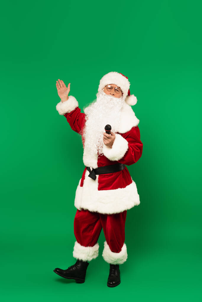 Санта-Клаус в костюме держит микрофон и указывает на зеленый фон - Фото, изображение