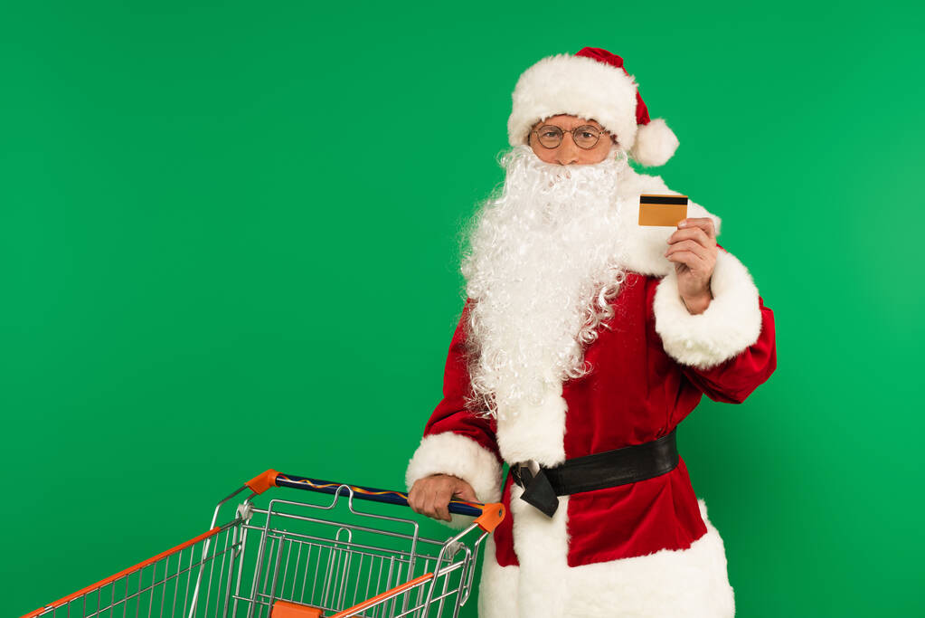 Santa Claus trzyma kartę kredytową w pobliżu koszyka na zielonym tle - Zdjęcie, obraz