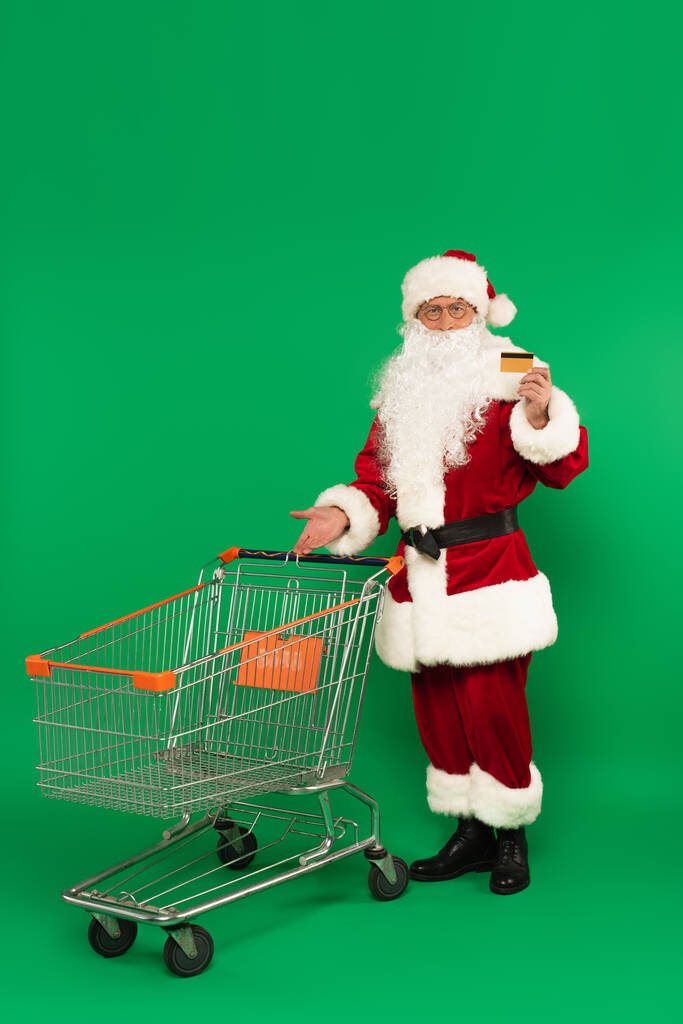 Święty Mikołaj trzyma kartę kredytową i wskazuje na koszyk na zielonym tle - Zdjęcie, obraz